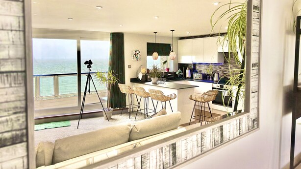 A modern kitchen with a large island, dining area with wicker chairs, and a view of the sea. - Stunning Sea View Two Bed Apartment - With Balcony (Southbourne)