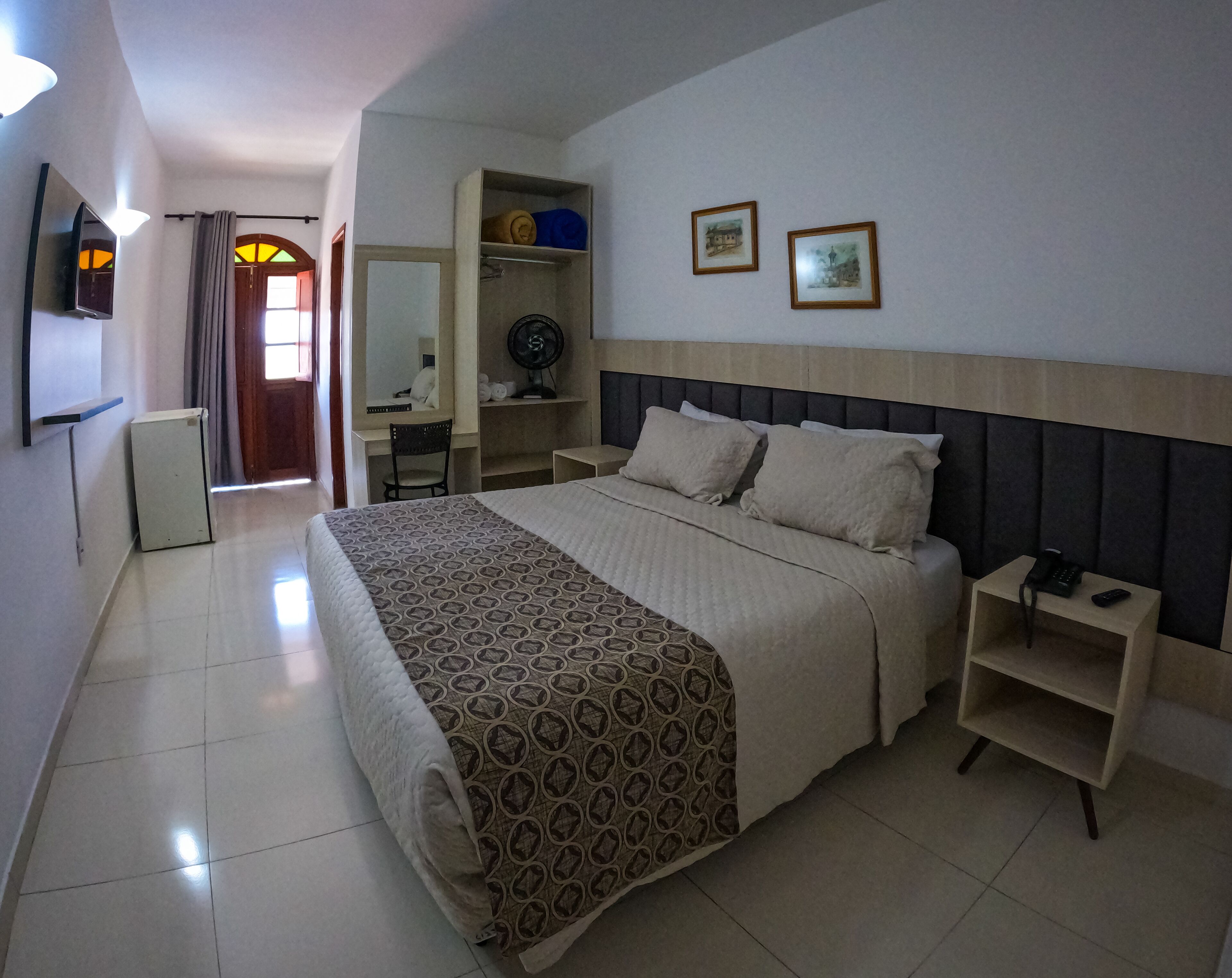 Una habitación de hotel con una cama grande, un escritorio con silla, un armario y un pequeño ventilador.