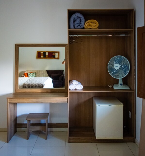 A compact room with a wooden desk, a small refrigerator, a fan, and a mirror reflecting a bed with pillows.
