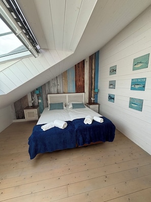 A bedroom with a sloped ceiling, a bed with blue bedding, white towels, and nautical-themed wall art.