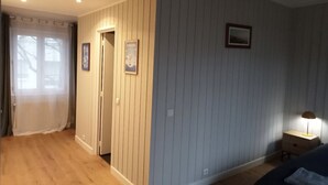 Une chambre avec un plancher en bois, un lit, une table de chevet avec une lampe et une porte donnant sur une autre pièce.