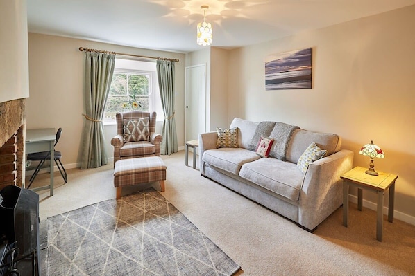 A cozy living room with a sofa, a patterned rug, a small table with a lamp, a chair, and a view of the outdoors through a window. - Host & Stay | Melody's Cottage (Berwick-upon-Tweed)