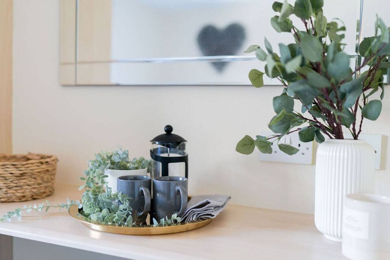 Un taulell de cuina amb una cafetera, dues tasses, un safata amb plantes i un vas blanc amb fulles verdes.