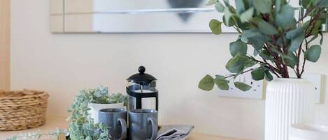 Un taulell de cuina amb una cafetera, dues tasses, un safata amb plantes i un vas blanc amb fulles verdes.