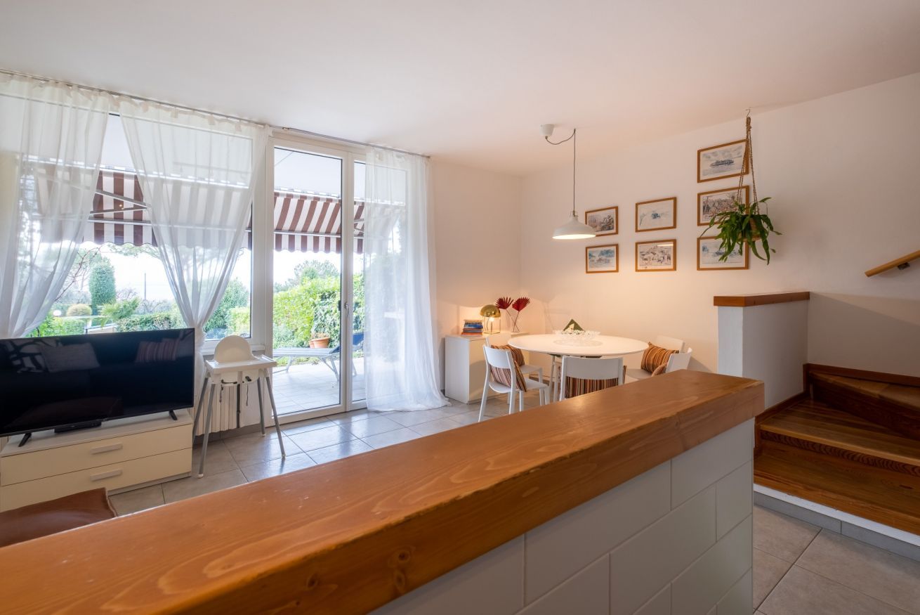Una cocina moderna con encimera de madera, un comedor con mesa y sillas blancas, un televisor y una puerta corrediza de vidrio que da a un balcón.