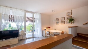 Une cuisine moderne avec un plan de travail en bois, un coin repas avec une table et des chaises blanches, un téléviseur et une porte-fenêtre coulissante donnant sur un balcon.