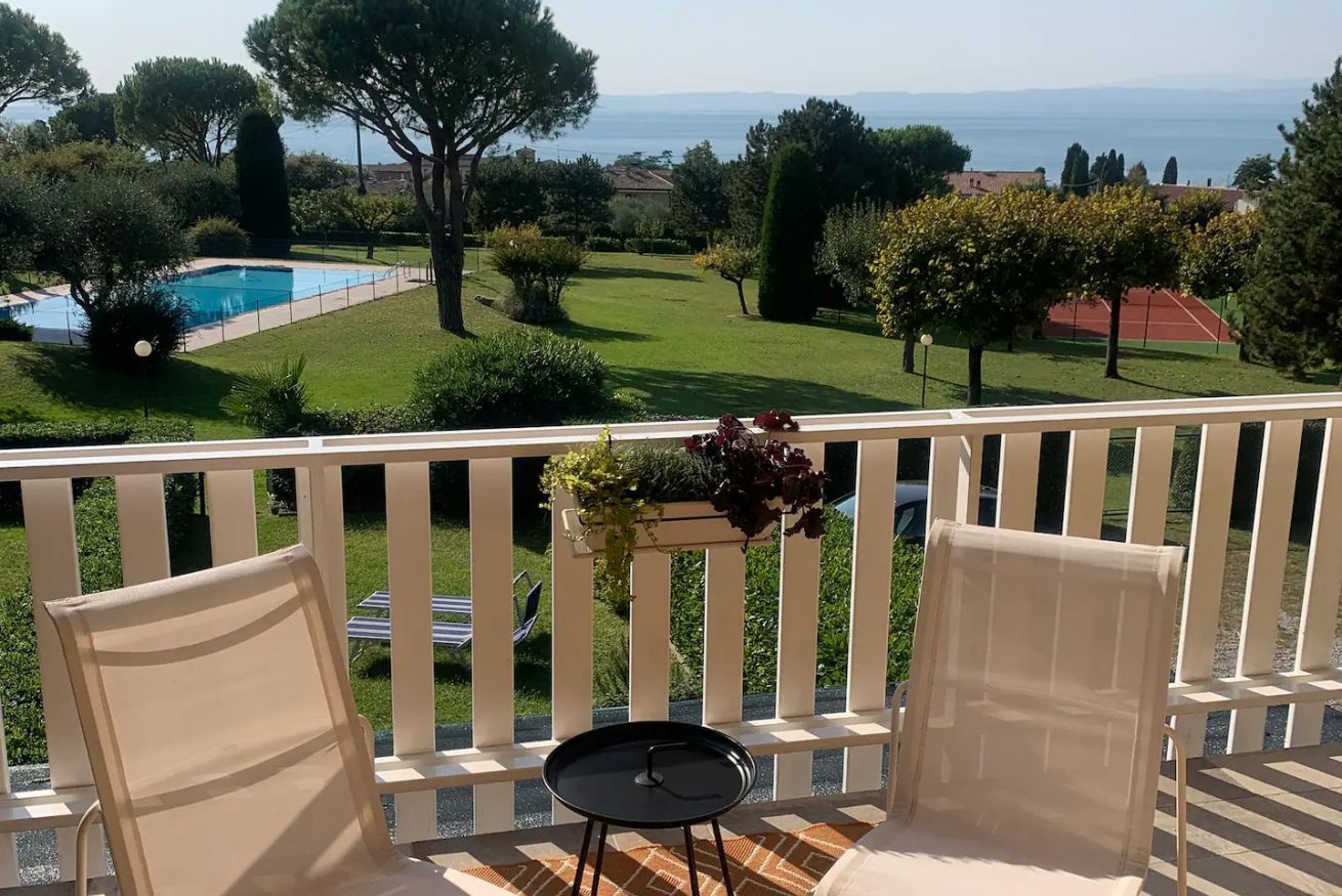 A balcony with two chairs and a small table, overlooking a swimming pool and a green lawn with trees and a tennis court.
