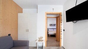 Een modern interieur met een grijze bank, een klein wit tafeltje met decoratieve objecten, een flatscreen televisie aan de muur en een houten deur die leidt naar een slaapkamer met een bed en een wandplank.