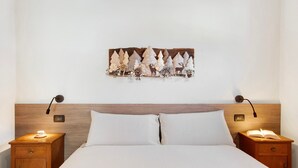 Een net opgemaakt bed met witte lakens, twee nachtkastjes met lampen, en een wanddecoratie van bomen en bloemen.