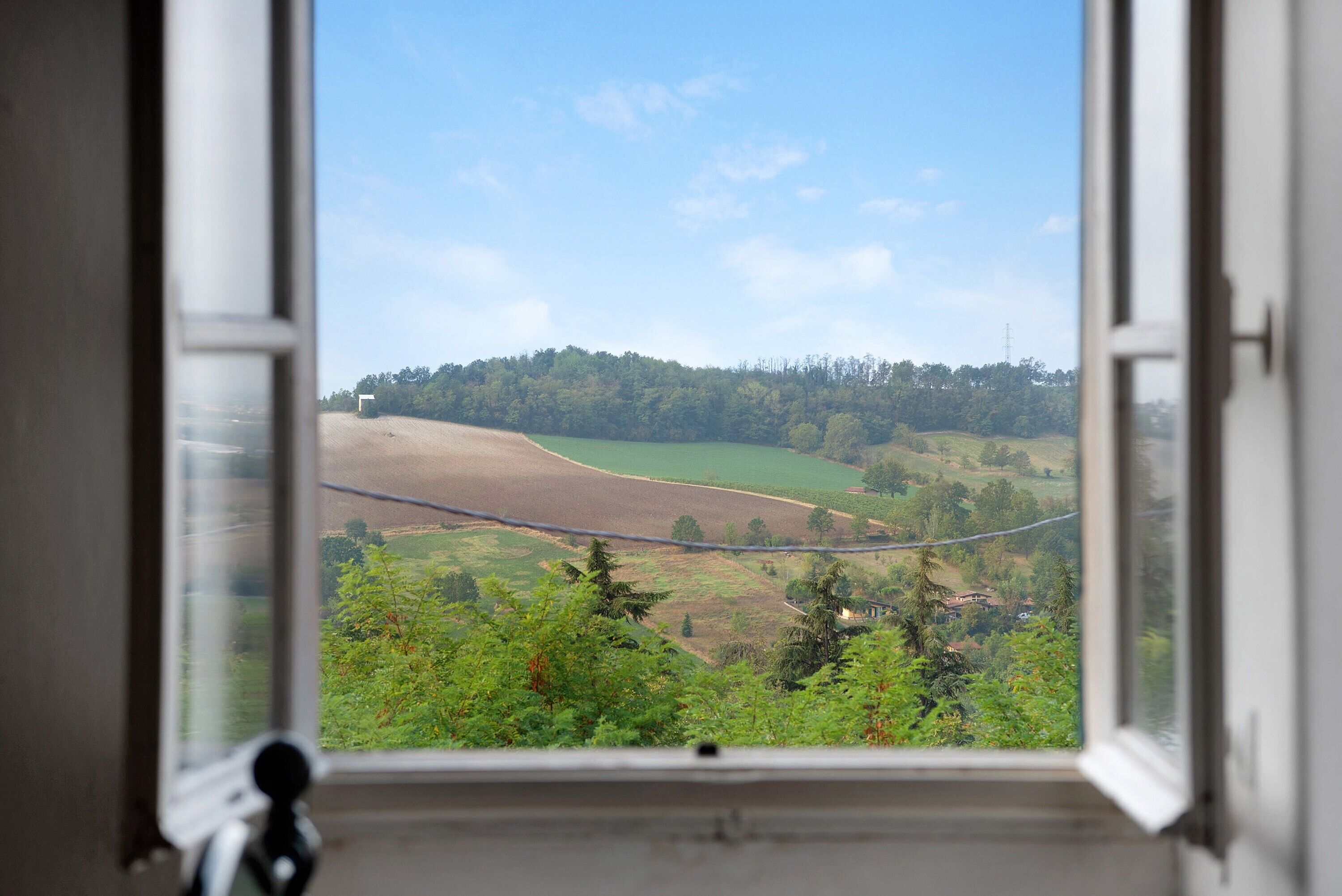 Blick durch ein offenes Fenster auf eine ländliche Landschaft mit Feldern, Bäumen und einer entfernten Hügelkette.