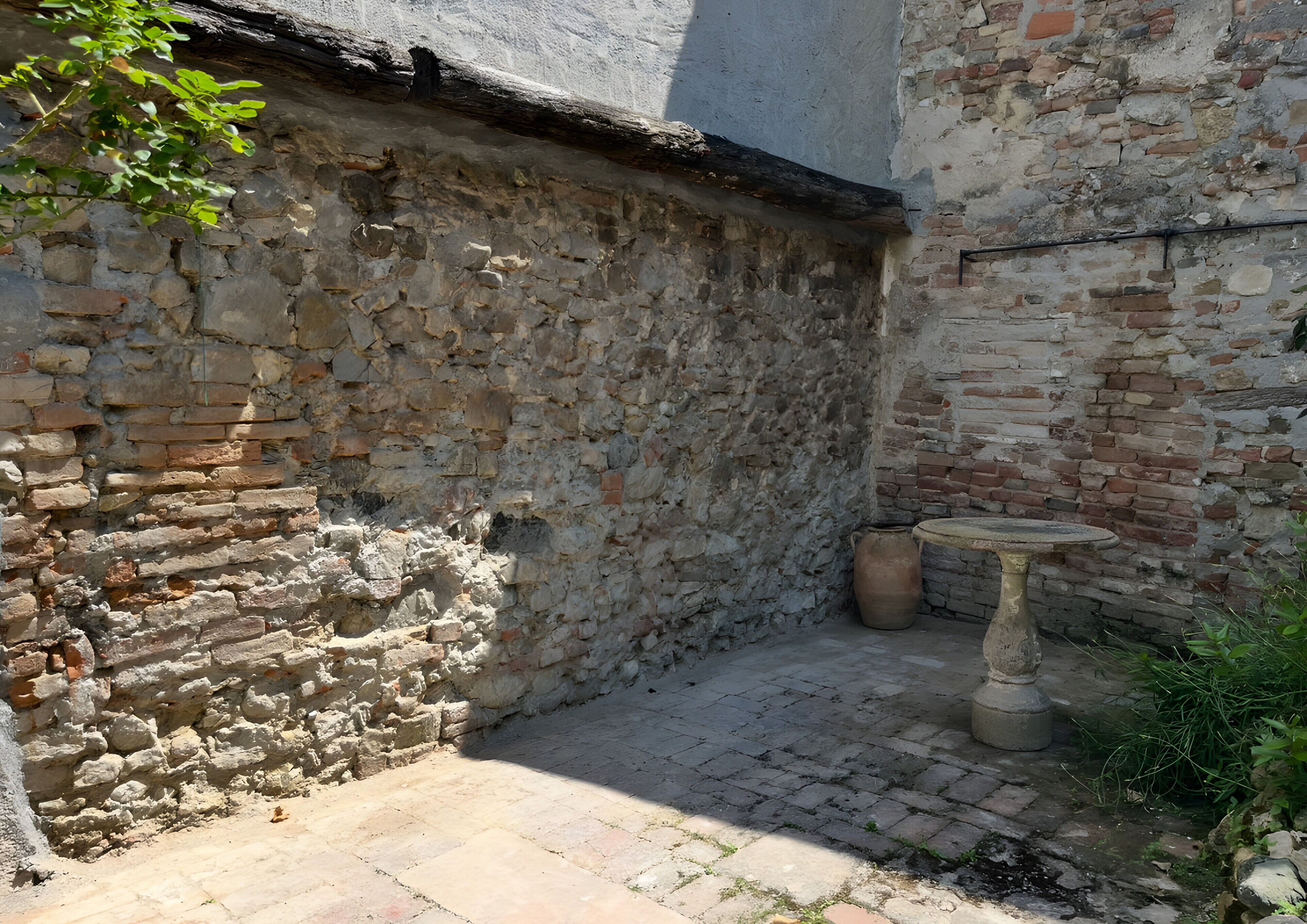Un muro de piedra con una superficie rústica e irregular, una mesa redonda pequeña y una planta en maceta en un espacio exterior sombreado.
