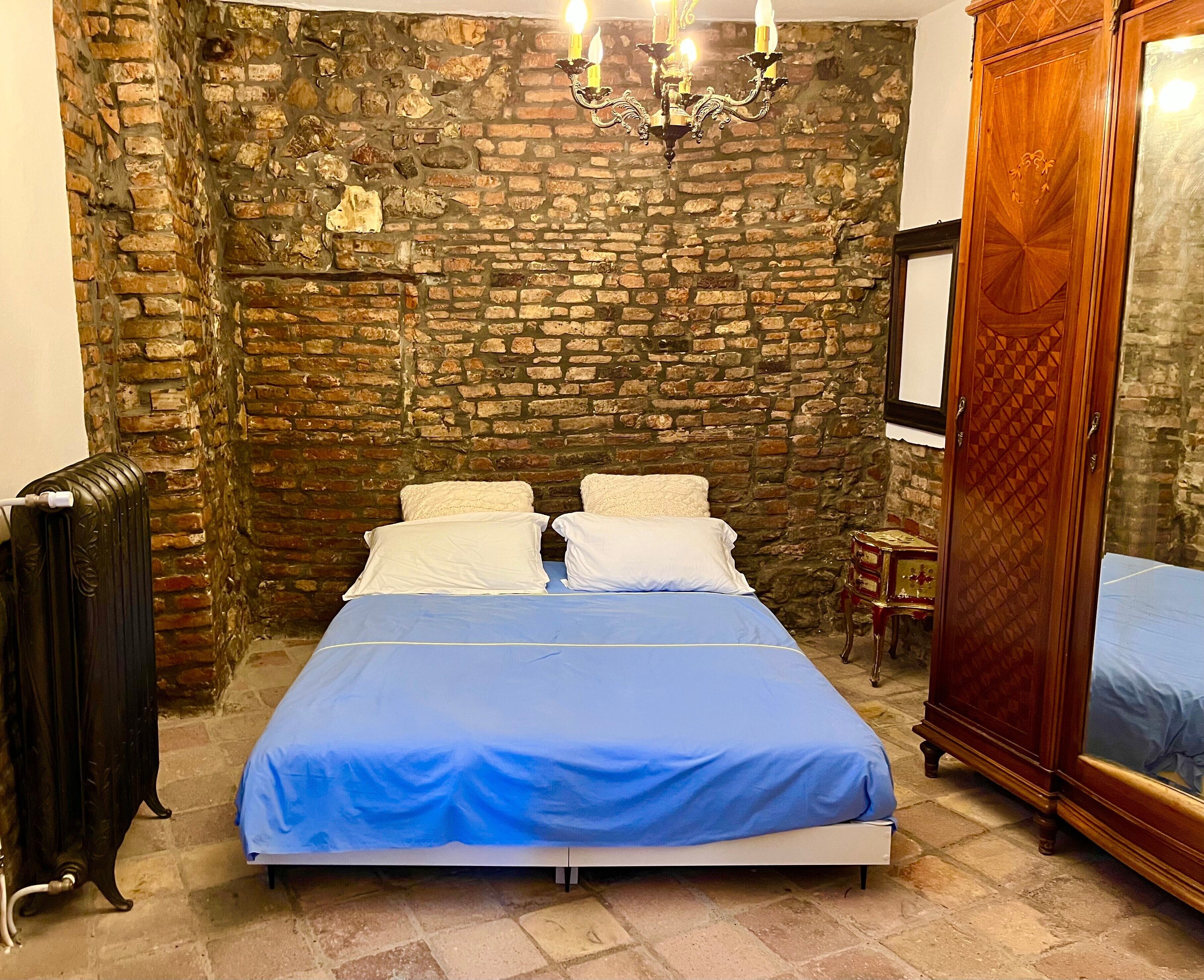 Een slaapkamer met een bakstenen muur, een bed met blauw beddengoed, een houten garderobe en een radiator.