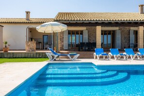 Ein Pool mit Liegestühlen und Sonnenschirm, eine Steinsäule und ein Gebäude mit Ziegeldach und Fensterläden.