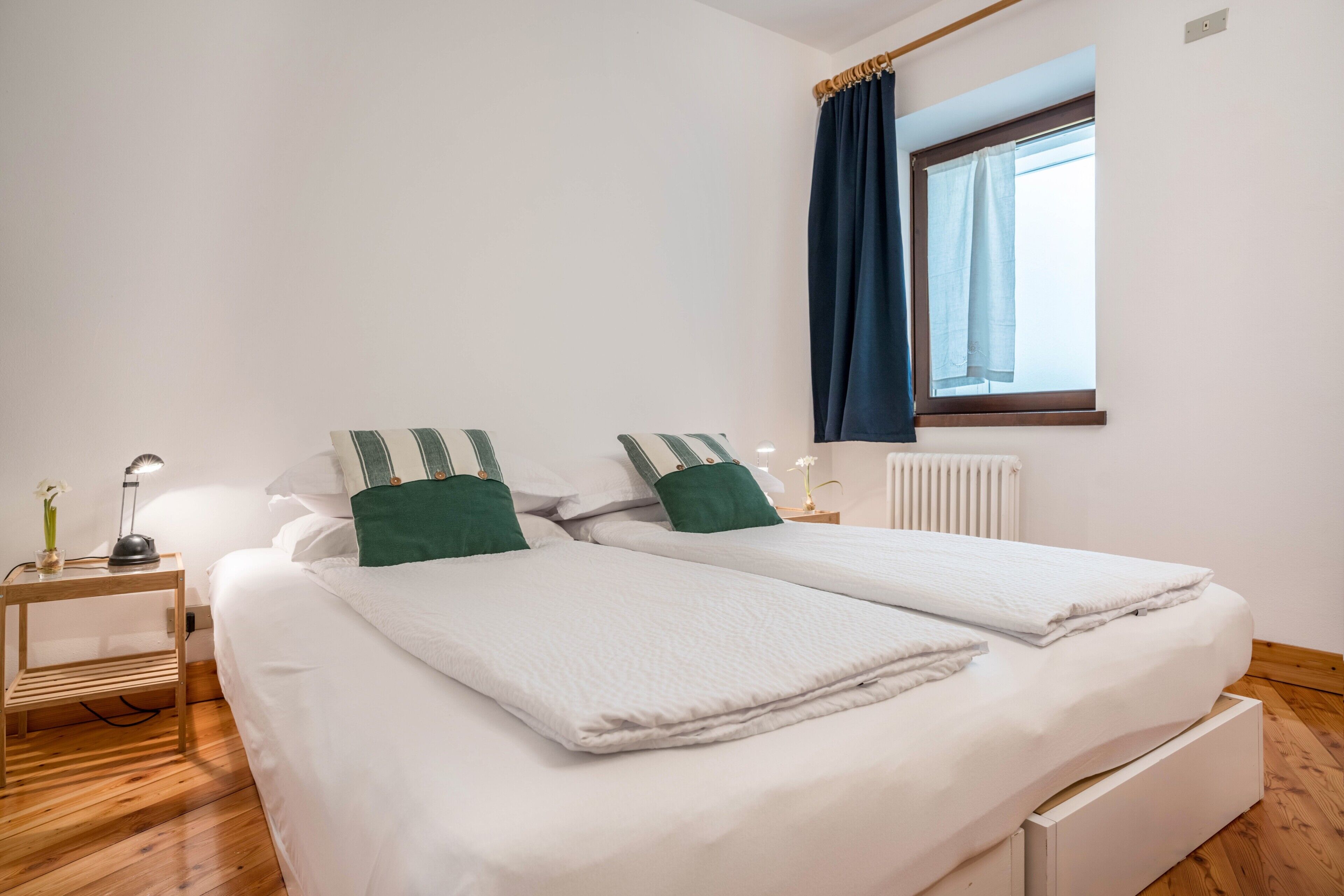 A double bed with white bedding and green pillows, a bedside table with a lamp, and a window with blue curtains.