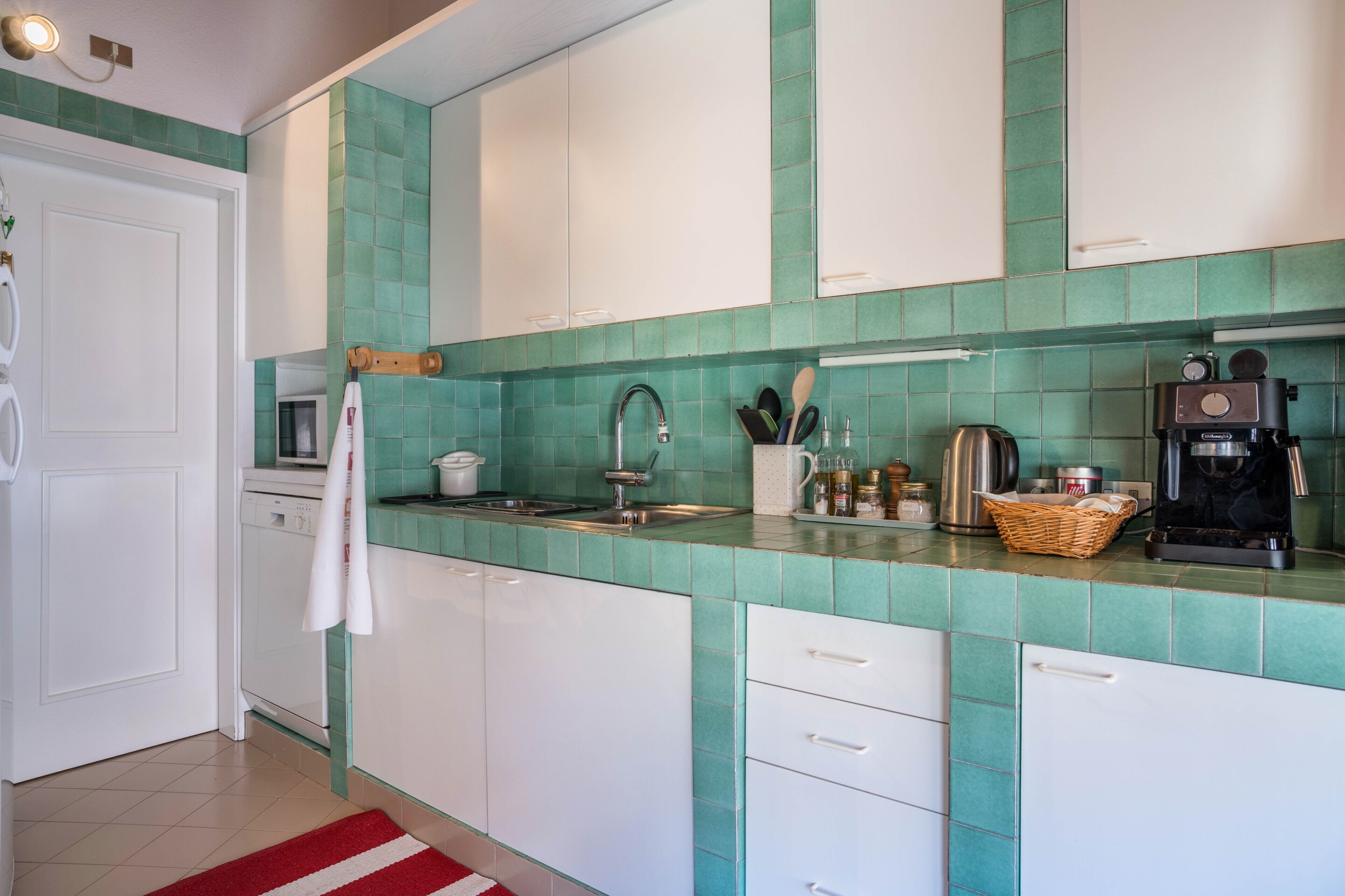Una cocina con baldosas verde mar, armarios blancos, una cafetera y un tostador.