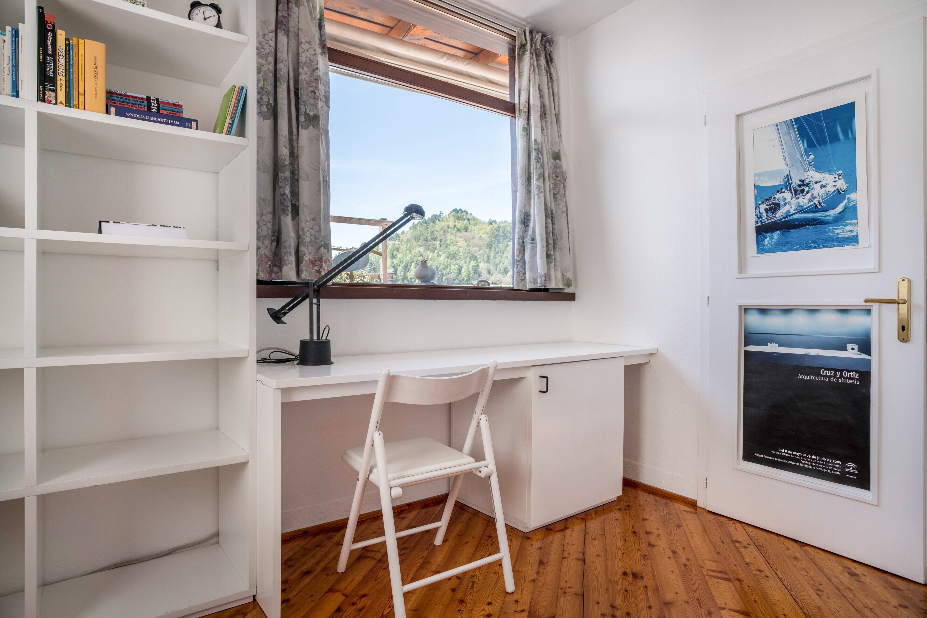 A room with a white desk, a white chair, a bookshelf, a window with curtains, and a framed picture on the wall.