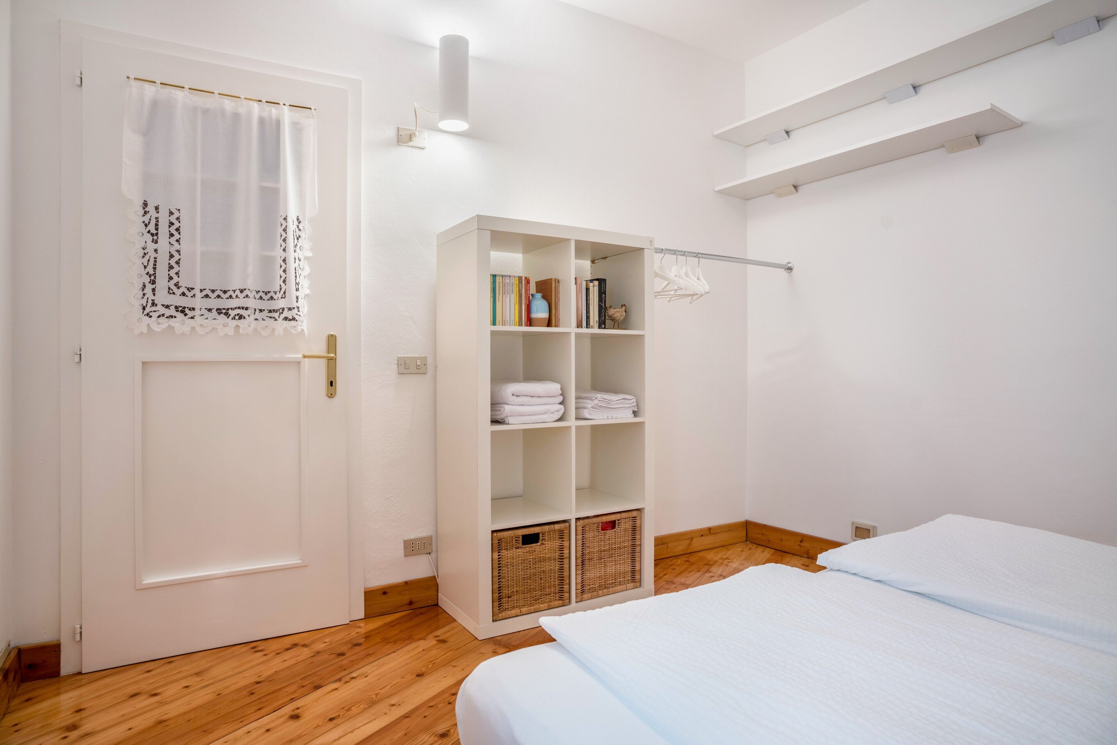 Une chambre avec un plancher en bois, une armoire blanche avec des étagères, un lit avec une literie blanche et une porte blanche avec un rideau.
