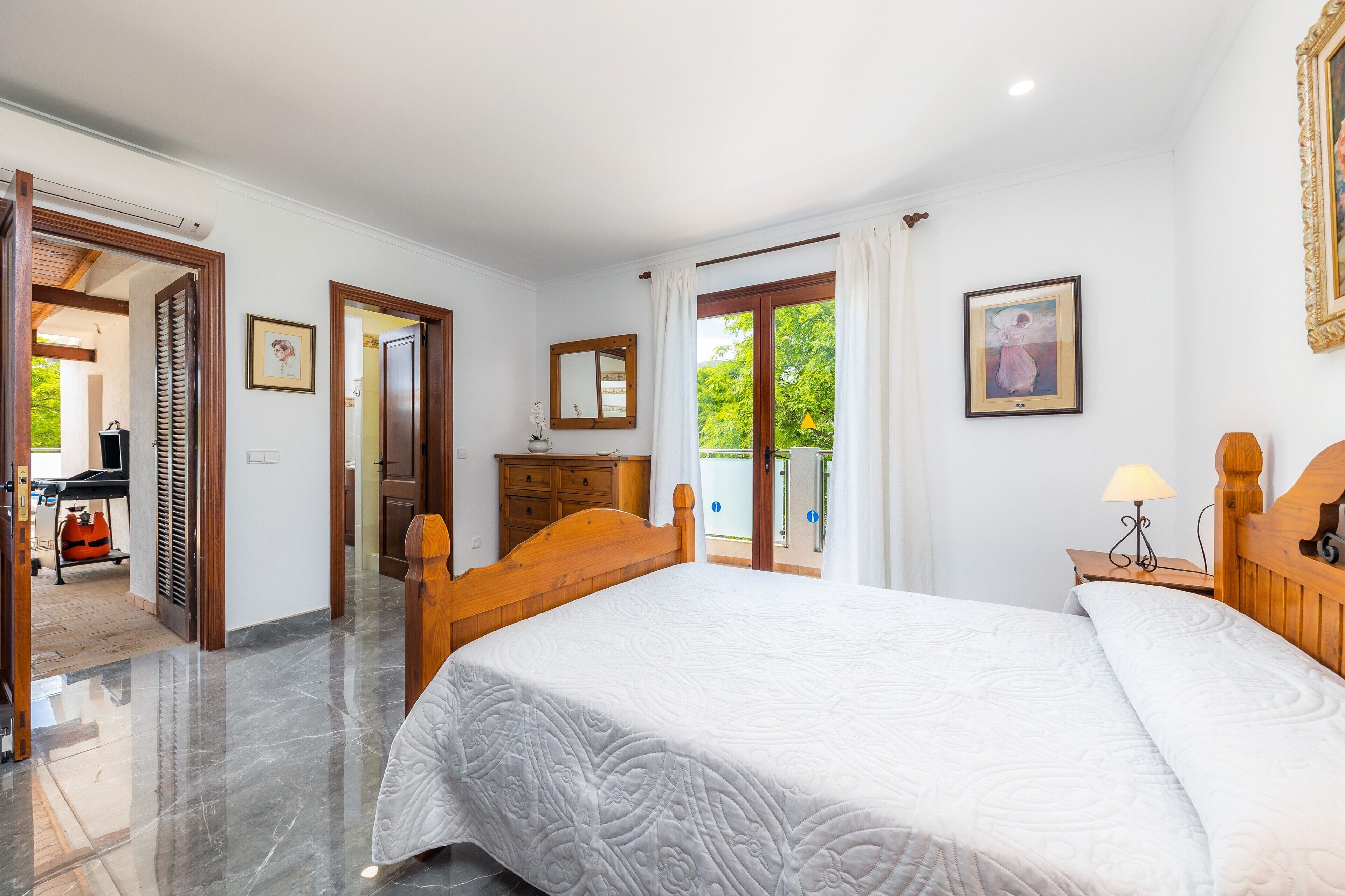 A bedroom with a large bed, wooden headboard, a nightstand with a lamp, a dresser, and a door leading to a balcony.