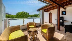 A balcony with wicker furniture, a grill, and a view of a body of water and greenery. - Apartment 'Cala' with Mountain View, Wi-Fi and Air Conditioning (puerto pollensa)