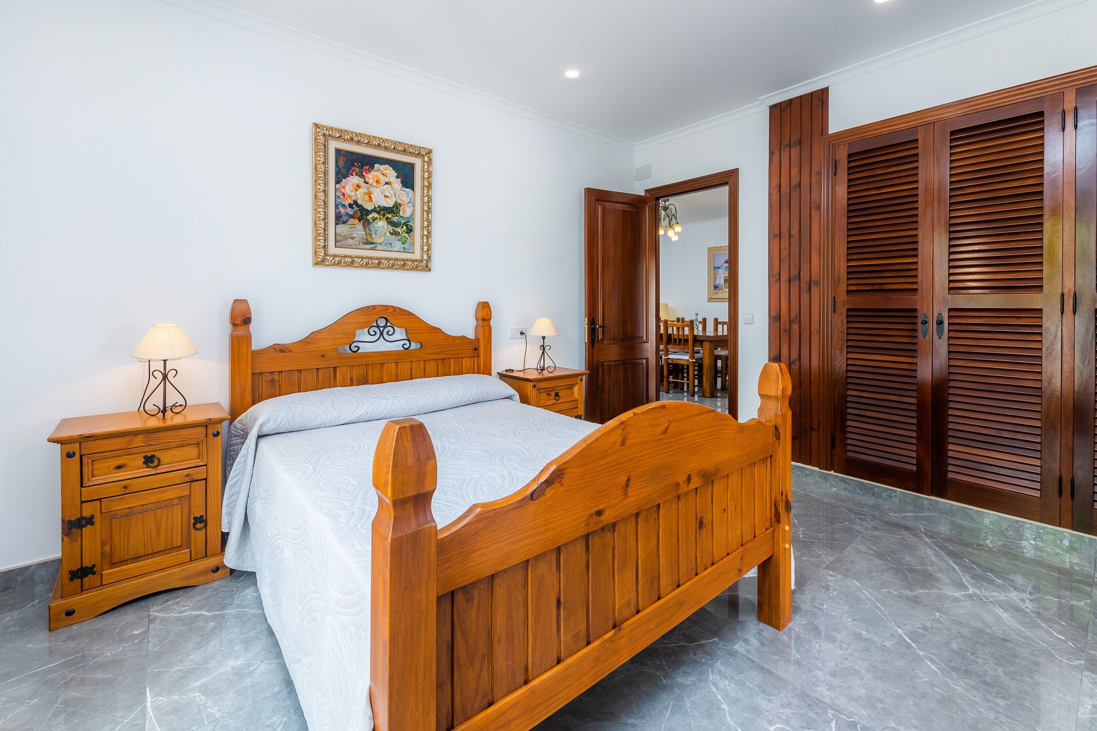 A bedroom with a wooden bed, a nightstand, a bedside table, a framed picture on the wall, and a wooden wardrobe.