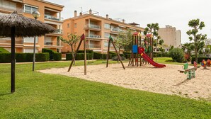 Um parquinho com balanços e escorrega, cercado por grama bem cuidada e sebes, com prédios de apartamentos ao fundo.