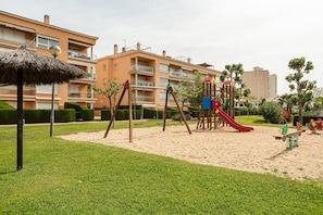 A playground with swings and a slide, surrounded by well-maintained grass and hedges, with apartment buildings in the background.