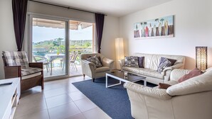 Sala de estar com sofá, poltrona, mesa de centro e vista para uma piscina e vegetação através de uma porta de vidro.