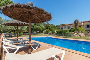 A swimming pool with lounge chairs and thatched umbrellas, surrounded by a fenced area with a building in the background.