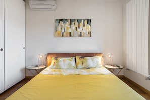 A bedroom with a bed, two bedside tables with lamps, a wall-mounted air conditioner, and a painting.