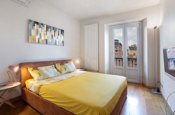 A bedroom with a bed, bedside table, lamp, artwork, window, and radiator.