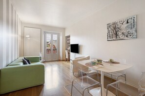 A modern living room with a green sofa, a dining table with chairs, a flat-screen TV mounted on the wall, and a cityscape artwork.