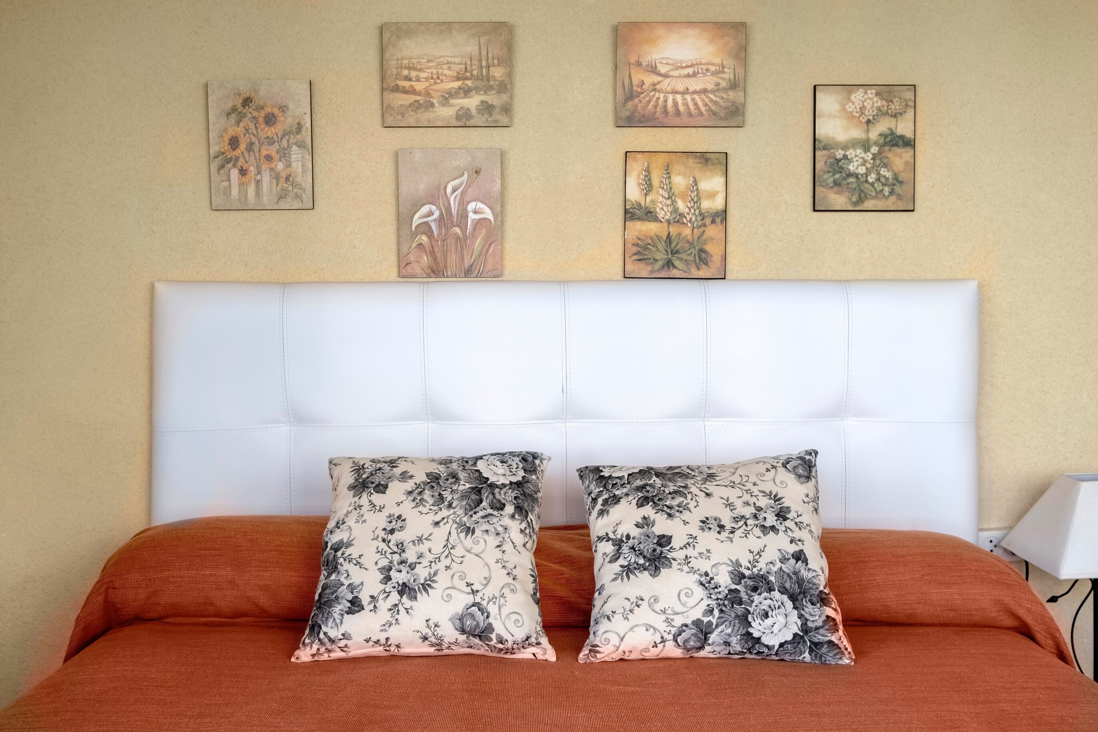 Una cama con cabecera blanca, dos almohadas estampadas con motivos florales y cuatro cuadros en la pared.
