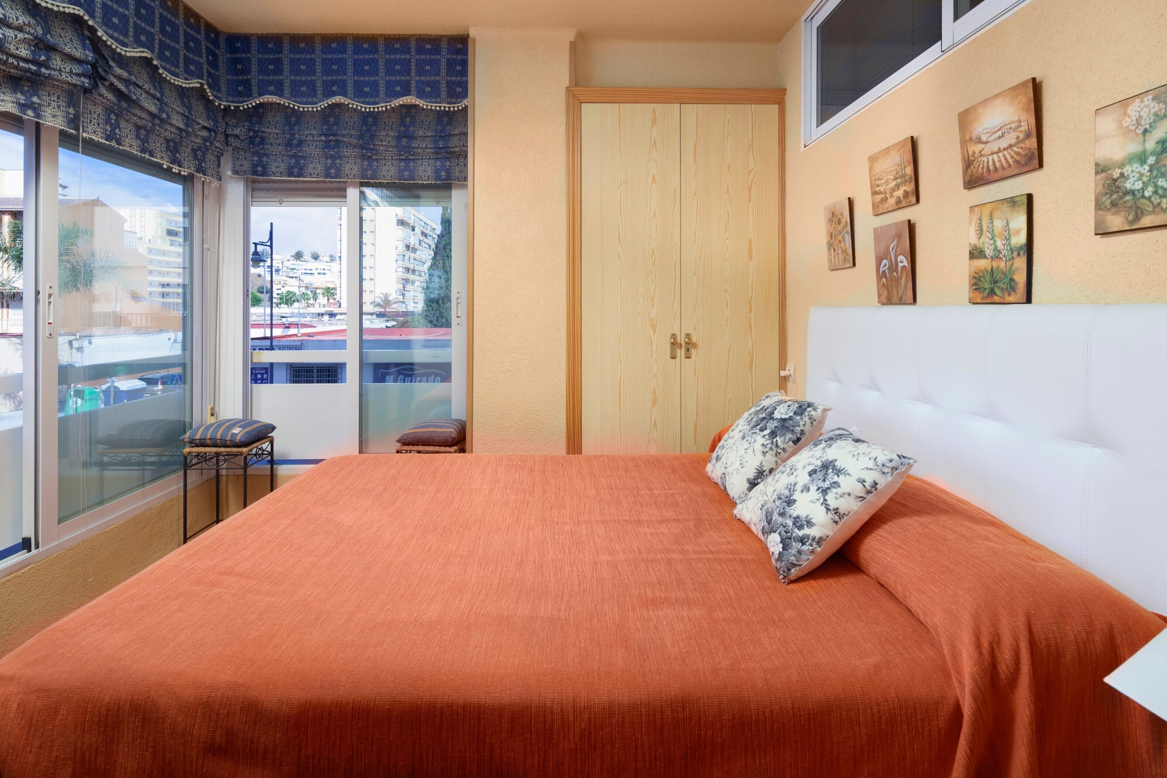 Un dormitorio con una cama grande, un armario de madera, una mesita pequeña y una ventana con vistas a edificios.