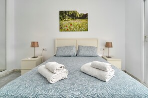 Een slaapkamer met een bed, twee nachtkastjes, twee lampen, een aan de muur hangend schilderij en twee opgerolde handdoeken op het bed.
