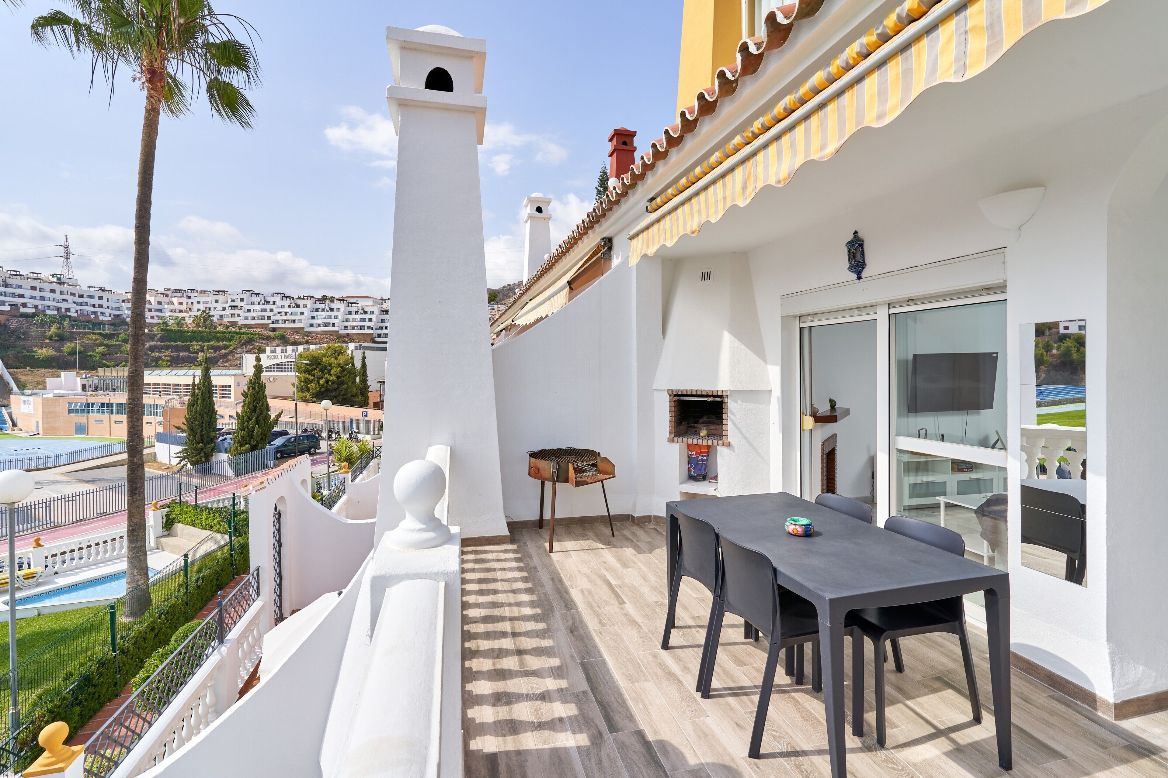Un balcón con mesa y sillas, una parrilla y vistas a una piscina y edificios.