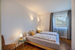 Un dormitorio con cama, una silla, una mesita de noche, una lámpara y una ventana con cortinas.