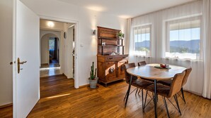 A dining area with a wooden table, chairs, a cabinet, and a view through large windows. - Holiday Apartment 'Weitblick Kaltern' with Mountain View, Private Terrace, and Garden (Caldaro sulla strada del vino)