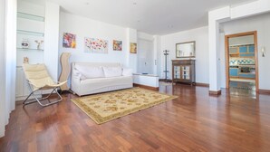Un salon spacieux avec un canapé blanc, un fauteuil beige, un tapis à motifs et un coin cuisine visible à travers une porte ouverte.