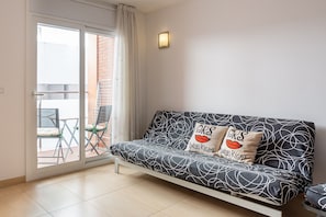 A modern living room with a sofa, two pillows, a wall-mounted light, and a sliding glass door leading to an outdoor area with a table and chairs.