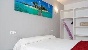 Un dormitorio con cama, pared blanca, un cuadro de playa y una estantería con toalla y tabla.