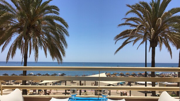Une table dressée avec une nappe bleue, des chaises blanches et un verre d’eau, donnant sur une plage bordée de palmiers et parsemée de parasols.