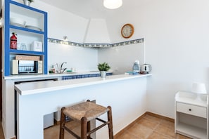 Una cocina con mostrador blanco, estanterías azules, una silla de mimbre, un armario blanco, un reloj y una planta en maceta.