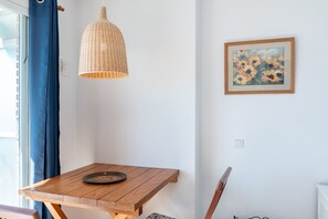 Una habitación con una mesa de madera, una lámpara colgante de mimbre, un cuadro floral enmarco y una cortina azul.
