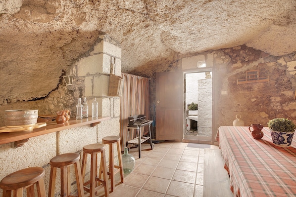 A rustic kitchen with a stone wall, wooden bar stools, a tiled floor, and a view through an open door.
