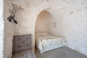 Une petite pièce aux murs de pierre, avec un lit simple, un banc rembourré et une tête de cerf accrochée au mur.