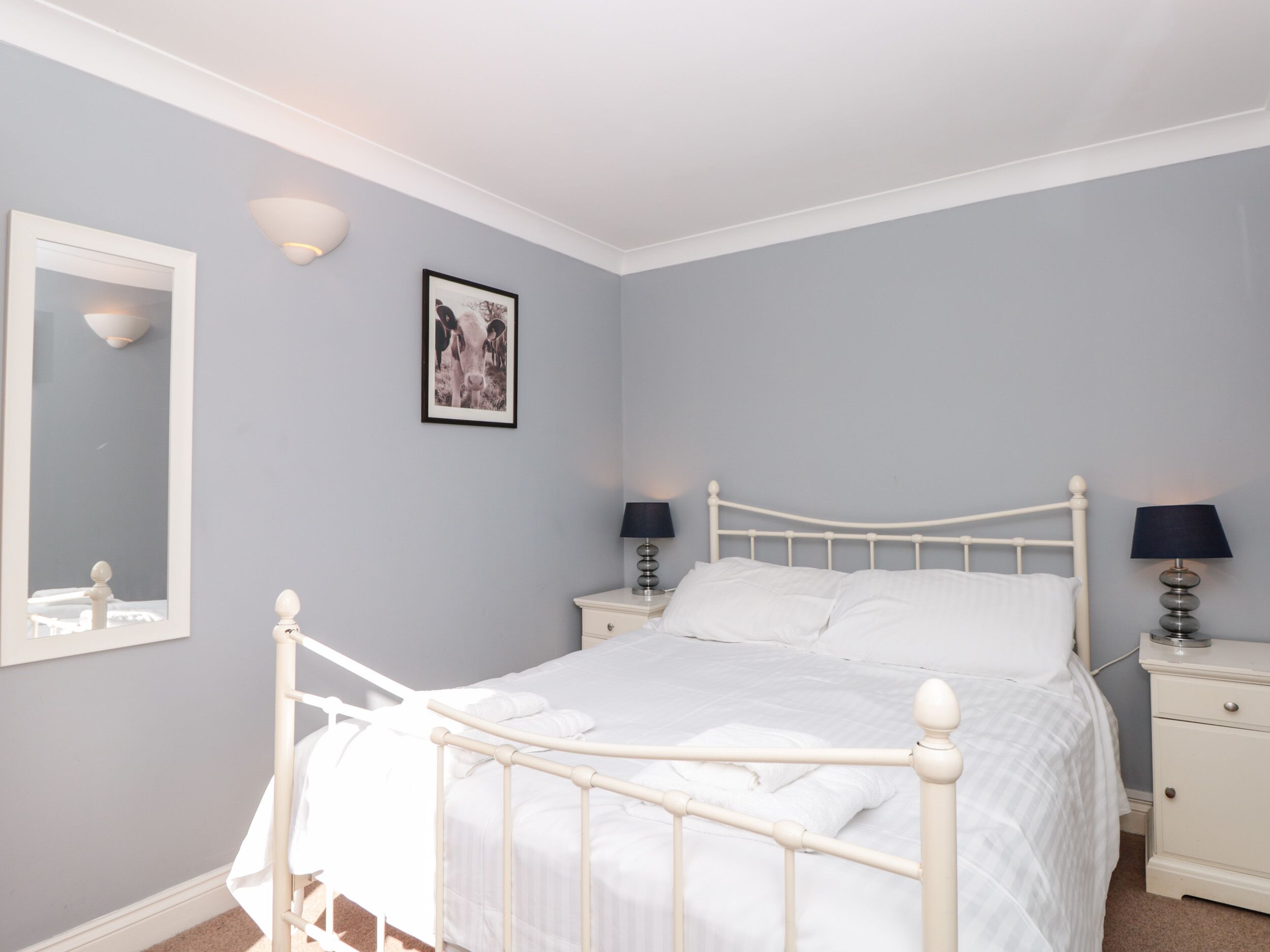 Un dormitorio con una estructura de cama metálica blanca, dos mesitas de noche con lámparas, un espejo y un cuadro enmarcado en la pared.
