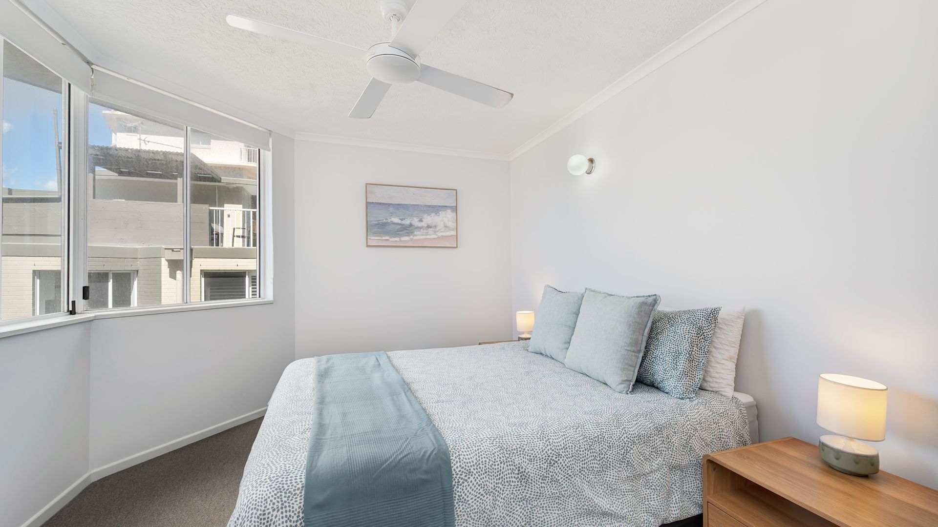 A bedroom with a bed, a ceiling fan, a window, a painting, and a bedside table with a lamp.