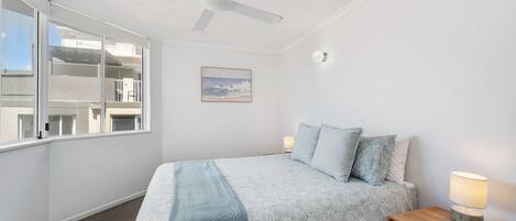 A bedroom with a bed, a ceiling fan, a window, a painting, and a bedside table with a lamp.