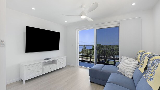 Een moderne woonkamer met een flatscreen televisie, een witte tv-kast, een blauwe bank en een balkon met uitzicht op zee.