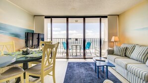 A balcony with a view of the sea, a dining area with yellow chairs, a striped sofa, and a TV on a table. - 0910 Gone To The Beach by Atlantic Towers (Carolina Beach)